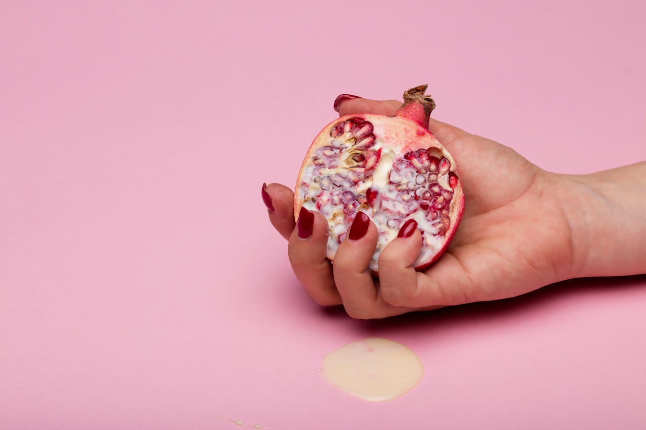 gallery-2 Close-up of a hand with painted nails holding a sliced pomegranate on a pink background.