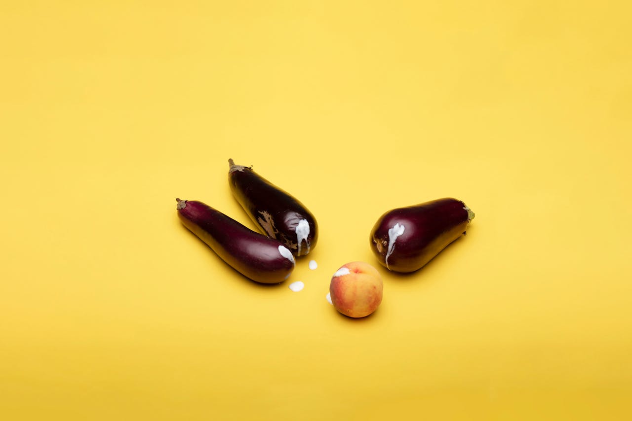 gallery-1 Playful composition with eggplants and peach on a vibrant yellow background.